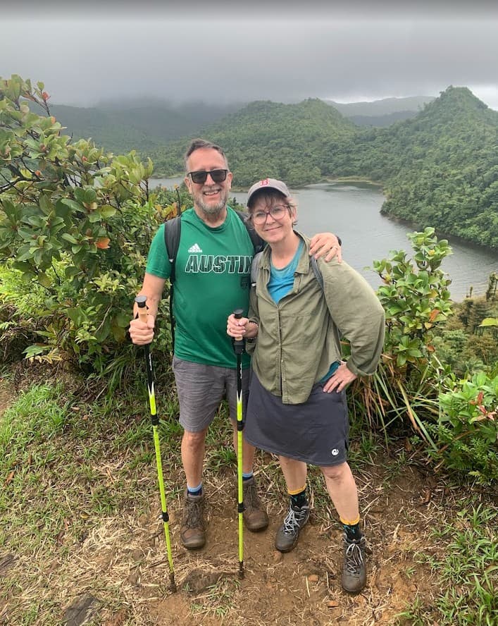 A smiling man and woman on a hiking trail overlooking lush green mountains and a lake in Dominica